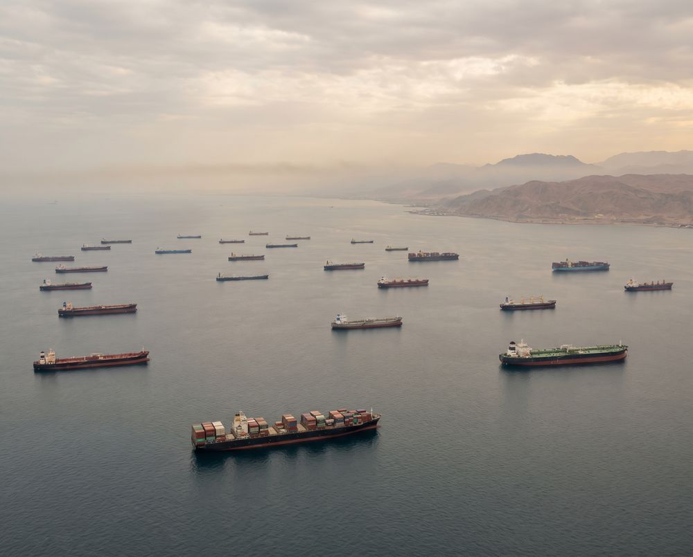 Aerial view of tanker and container ships anchored in Gulf waters during the Hormuz strait closure — China-India shipping route extended by 14-21 days via Cape of Good Hope.