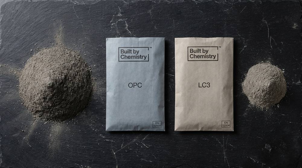 Two branded Built by Chemistry cement bags side by side on a black slate surface, with a large dense mound of grey ash beside the OPC bag and a smaller mound beside the LC3 bag, representing the relative CO2 footprint of each.