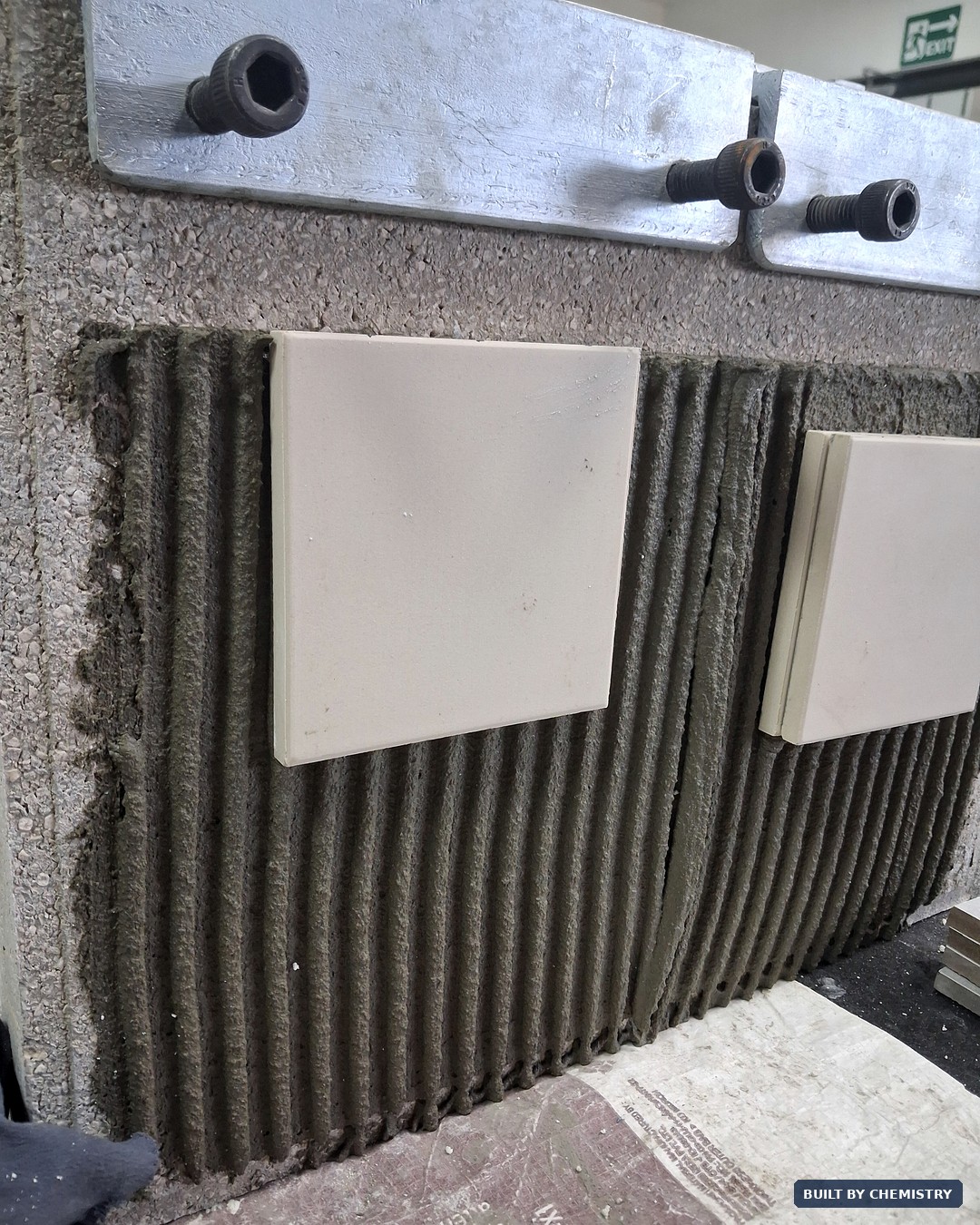 EN 1308 slip test on a vertical board — two white ceramic tiles bedded onto combed vertical ridges of grey cementitious tile adhesive, held under the aluminium restraint bar to measure how far each tile slides downward over time.